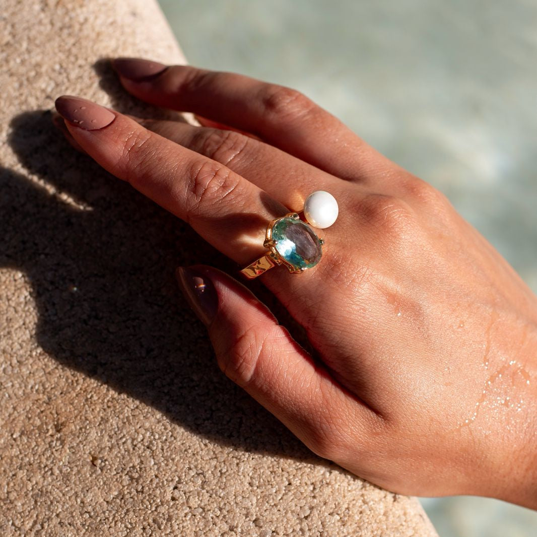 Hand wearing a ring with a green gemstone and pearl on a sandy surface.