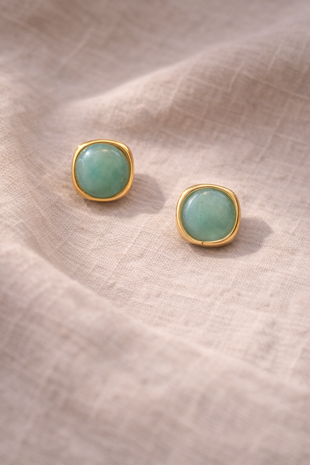 Pair of gold-framed green gemstone earrings on a beige fabric background
