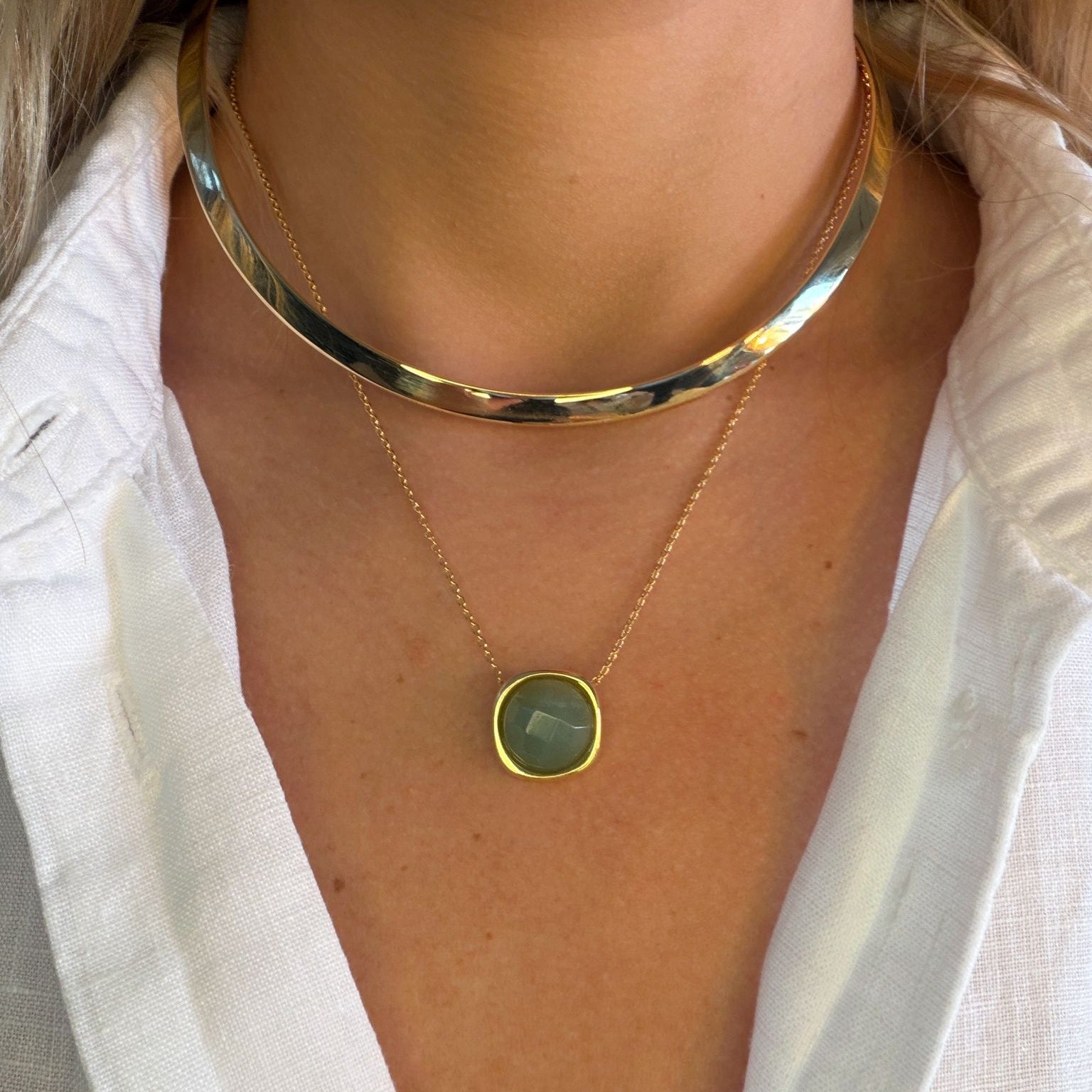Gold collar and gold necklace with a green pendant worn by a person with a linen shirt